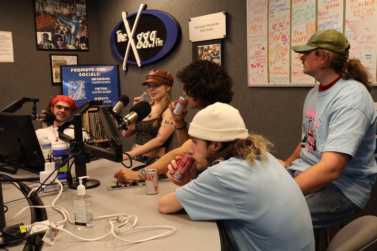 The band around the table in the WMCX studio between takes, mics overhead and the station's whiteboards covering the back wall